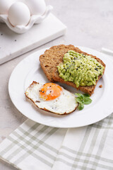 Slices of gluten-free sunflower seeds bread with mashed avocado, fried egg and sesame seeds on white plate on green checkered napkin