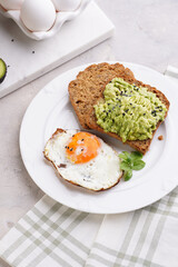 Slices of gluten-free sunflower seeds bread with mashed avocado, fried egg and sesame seeds on white plate on green checkered napkin