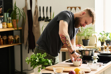Handsome young Caucasian man with tattoos on arms standing in kitchen crushing spices with mortar and pestle