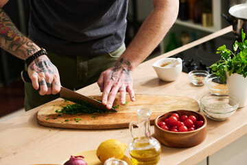Horizontal medium section shot of unrecognizable chef with tattooes on arms chopping fresh cilantro...