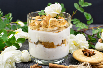 Multi-layered cottage cheese and yogurt dessert with banana and cookies in a glass glass and white roses on a black (dark) background.