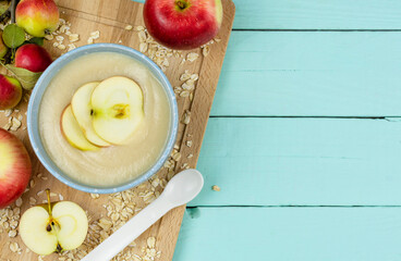 Oatmeal porridge for the baby from ground cereals in a blue bowl, red ripe apples on a wooden board on a wooden background. Space for text. Baby's first complementary food, baby nutrition.
