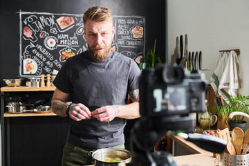 Stylish tattooed Caucasian man with beard on face filming content for food blog while cooking potatoes in saucepan