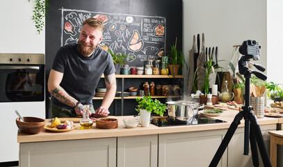 Joyful young adult Caucasian man with beard on face smiling at something while cooking dinner and filming process for blog