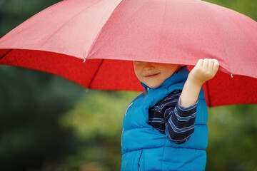 Happy child with an umbrella playing out in the rain in the summer outdoors. The boy smiles and hides his face under an umbrella