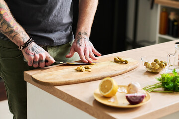 Unrecognizable tattooed man cutting pickled olives on wooden board in loft kitchen, medium section shot