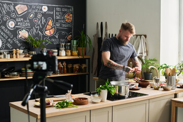 Handsome bearded food blogger standing in loft kitchen cooking dinner on camera peeling shrimps