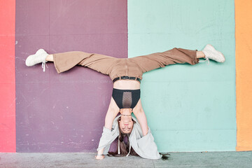 Full length powerful woman in casual wear performing handstand with split near colorful wall