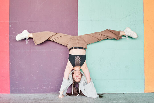 Full length powerful woman in casual wear performing handstand with split near colorful wall