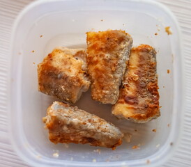 Fried pieces of pollock fish in a white plastic container close-up on the background of a light kitchen table