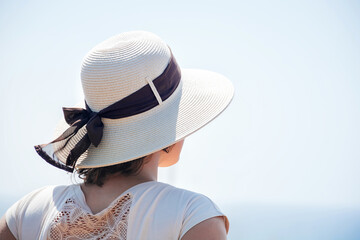 Back view soft focus on a young girl standing in a hat on her head from the sun with a ribbon with a bow. Woman looks at the sea on a hot sunny day. Tourist vacation in quiet solitude. Copy space.