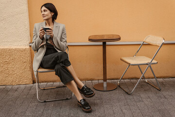 Full length caucasian adult woman sits at table in cafe drinking coffee outdoors. Brunette in grey...