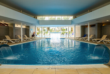 Water falling in swimming pool. Deck chairs arranged in room at luxurious resort. View of indoor...