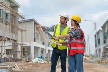Fototapeta na wymiar Asian female civil engineer and caucasian male architect wears safety vest with helmet discuss and look at blueprints in housing estate construction site