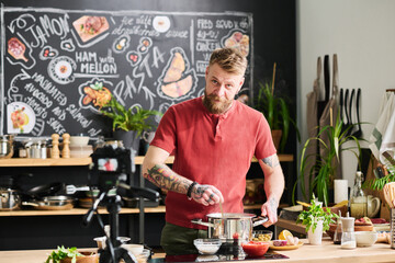 Handsome tattooed Caucasian man with beard on face standing in loft kitchen cooking pasta and...