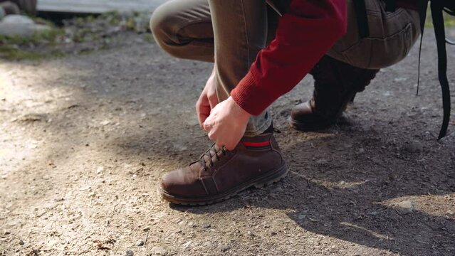 The guy ties the laces on brown boots standing on one knee on the ground.