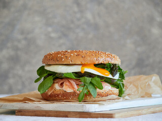 Front view of delicious bagel with salmon, cream cheese, arugula leaves and lamb's lettuce with egg yolk dripping. On wooden board and food wrap.