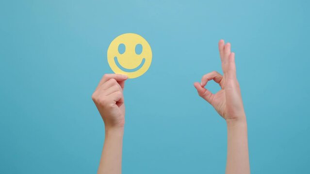 Female hand holding yellow happy smile face, good feedback rating and positive customer review, showing ok sign, satisfaction survey, mental health assessment, isolated on blue studio background wall