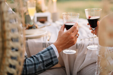 Hand of man and woman sitting on chairs  by a table and holding glasses with red wine. Outdoor wedding for two 