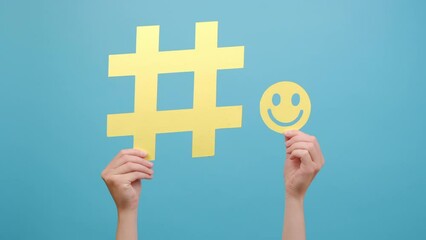 Close up of female hands holding big yellow hashtag sign and little happy smiling face icon, isolated on blue color background wall in studio with copy space for advertisement. Online and web concept