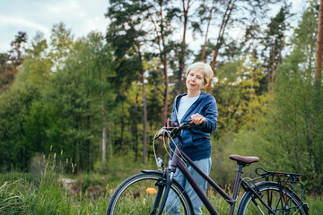 Fototapeta na wymiar Short blond hair aged woman enjoying healthy outdoor activities. Cycling. Smiling senior woman standing on a country lane with her bicycle.