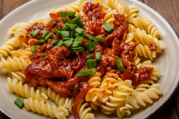 Beige plate with bolognese pasta on a wooden table sprinkled with green onions close-up.
