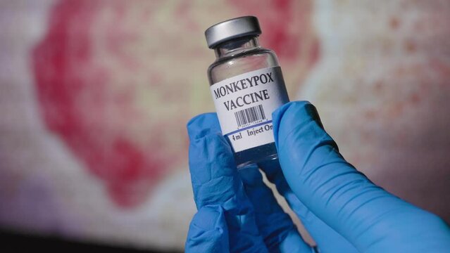 doctor holding a vial of monkeypox vaccine in front of a monitor with the monkeypox virus displayed