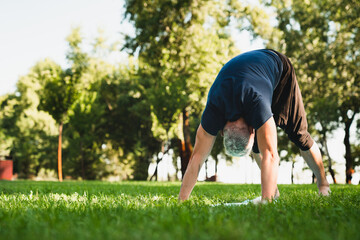 Caucasian mature fit healthy man in sporty clothes doing yoga exercises outdoors , standing in yoga position on green grass in public park