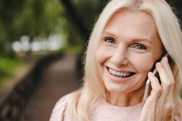 Happy mature caucasian woman talking on cellphone, calling to her friends, ordering food delivery, taxi on smart phone, looking at camera in park forest.