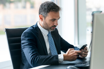 Elegant mature businessman using mobile phone while working with computer in his office in a modern startup.