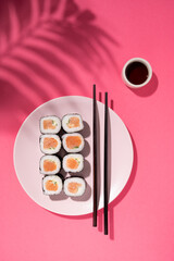 Japanese Sushi roll with salmon, cucumber and soy sauce with chopsticks in the plate on pink background with hard shadow palm leaf. Concept food photography. Top view and copy space