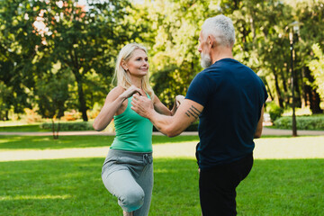 Mature fitness trainer coach instructor standing in balance helping woman practicing yoga, meditating and relaxing in park. Husband going in for sport with his wife.