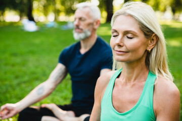 Close up portrait of mature caucasian couple meditating together on fitness mat in public park. Yoga class outdoors. Trainer coach sitting in yoga position with mature woman.