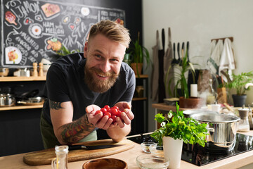 Cheerful young Caucasian man with beard on face and tattoos on arms holding cherry tomatoes in hands smiling at camera