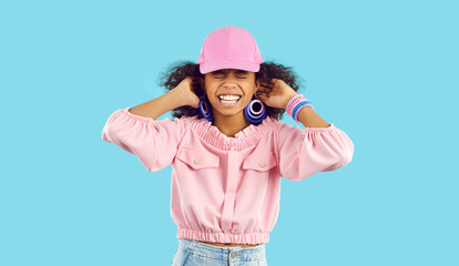 Portrait of happy cheerful child. Little ethnic kid standing in studio and laughing at funny joke. Cute Afro American girl puts on trendy pink ball cap, fixes her curly naughty hair and laughs happily - Powered by Adobe
