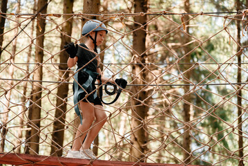 A little girl walks on a rope at a height with a belay under the open sky. The child has fun in the...