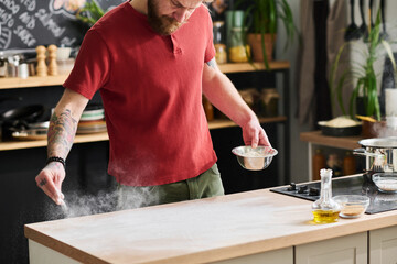 Modern professional chef in casual clothes dusting kitchen table with flour while cooking food to...