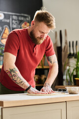 Bearded Caucasian man wearing casual outfit standing in loft kitchen kneading and flattening dough for pizza or pie