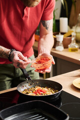 Vertical shot of unrecognizable man with tattooes on arms adding some tomato sauce to other ingredients in frying pan