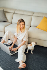 Happy blonde frelancer woman working from home. smiling and looking at small dog. Using computer laptop typing. Studing or online shopping Sitting on floor in sunny cozy living room lean on beige sofa