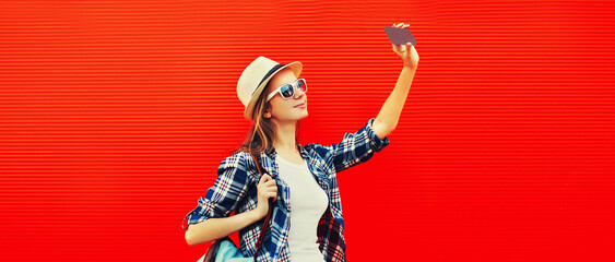 Portrait of happy young woman taking selfie with smartphone wearing summer straw hat, backpack on...