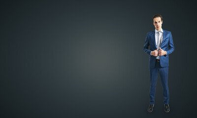 Young self-confident businessman in blue suit and grey tie isolated on dark grey wall background with blank place for your logo, mockup