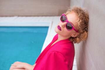 Fototapeta na wymiar portrait of marvelous stylish redhead curly young woman sitting in fuchsia jacket, stylish sunglasses and black underpants by the swimming pool. Fashion, style, leisure, recreation, travel, make up co