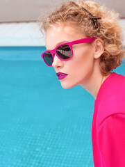 portrait of marvelous stylish redhead curly young woman sitting in fuchsia jacket, stylish sunglasses and black underpants by the swimming pool. Fashion, style, leisure, recreation, travel, make up co