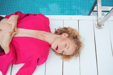portrait of marvelous stylish redhead curly young woman laying in fuchsia jacket by the swimming...