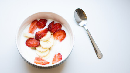 Natural greek white yogurt cream with fresh juicy strawberry and sliced banana in round bowl, spoon lying on white background. Top view. Healthy breakfast from nutritionist, good for digestion.