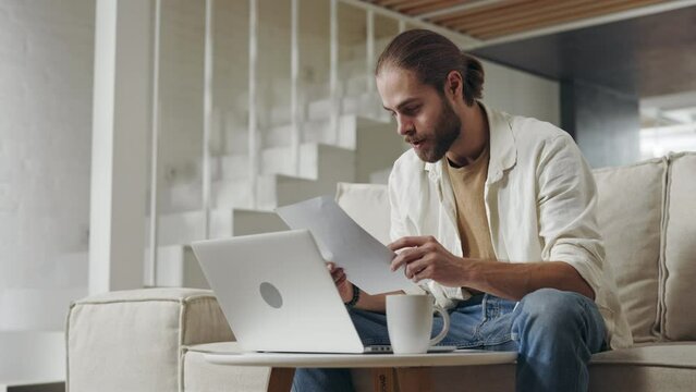 Handsome caucasian man doing paperwork while using laptop at home. Bearded guy in casual wear sitting on couch and calculating his expenses.