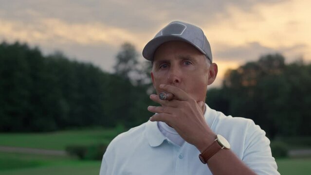 Senior golf player smoking cigar looking camera on sunset field course fairway.