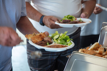 Two people (chest down) holding paper plates of BBQ meat, beans, salad, and garlic bread going through the buffet line
