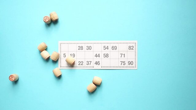 Many wooden barrels with numbers and a card for playing lotto or bingo. Board game. Gambling game with random element. Blue background. Flat lay. Top view.